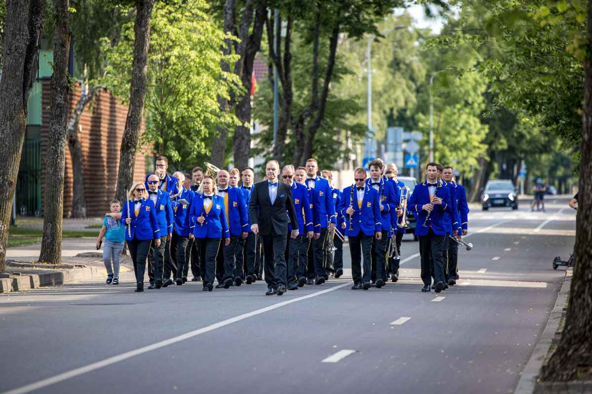 Muzikų grupė su mėlynais švarkais ir juodais kelnais žygiuoja tiesia gatve su medžiais šonuose